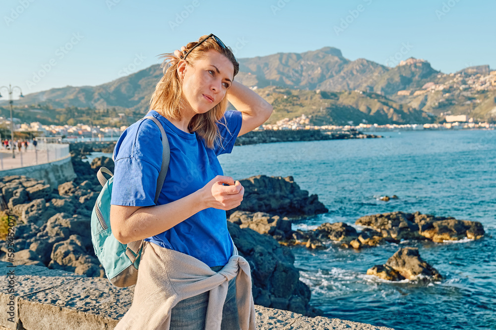Blond woman tourist with backpack looking into the distance, enjoying freedom by the sea. Female traveler relaxing in serene nature. Mental health, wellness, adventure travel and healthy lifestyle.