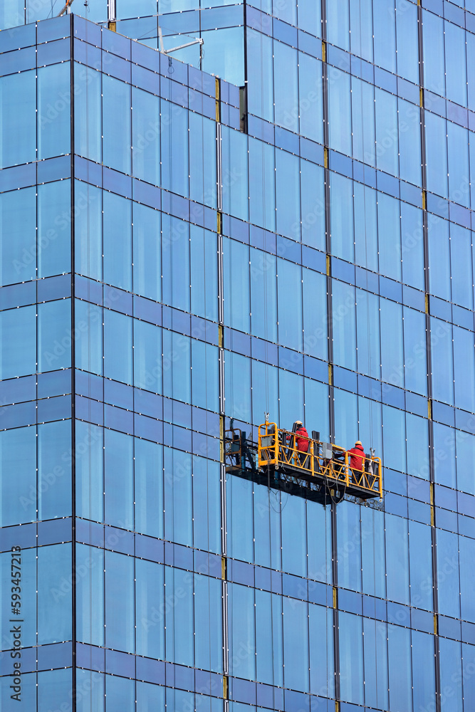 Builders in red overalls are mounting the glass facade of a multi-storey building on a suspended cradle.
