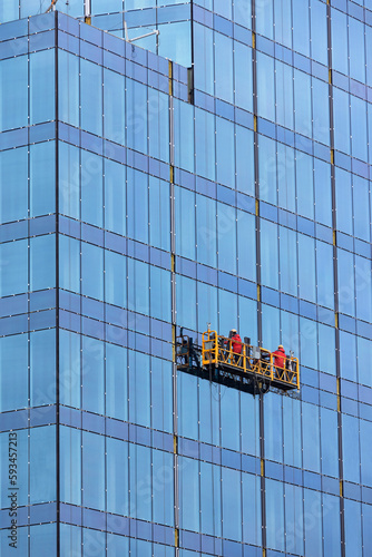 Obraz na płótnie Builders in red overalls are mounting the glass facade of a multi-storey building on a suspended cradle. w salonie