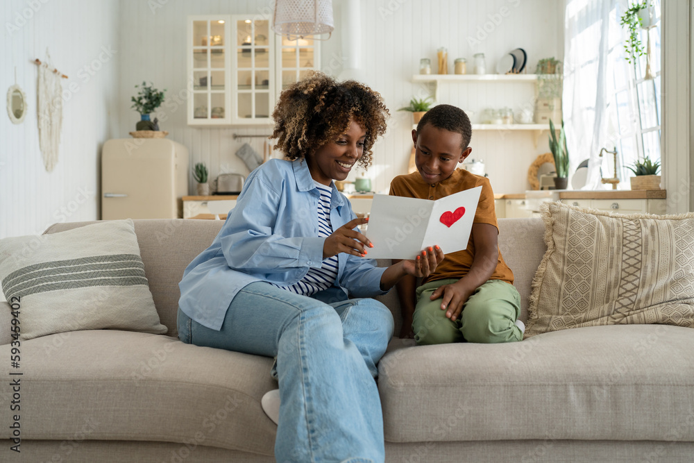 Caring African American son present mom greeting card for holiday event, mother day or birthday sit near mum reading postcard. Happy pleased young single mommy reading valentine together with child.