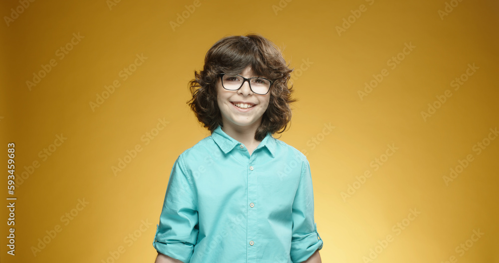 Little caucasian boy in blue shirt and glasses looking at camera, positively smiling, isolated on yellow background - emotions concept close up 