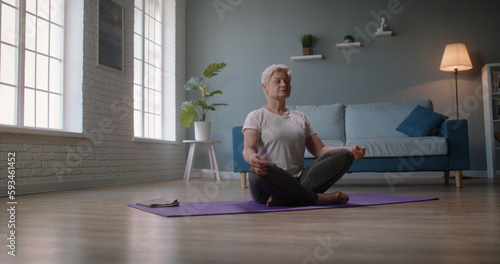 Mature caucasian woman doing yoga at home. Senior woman relaxing in lotus position on floor, keeping a healthy lifestyle - wellness concept 