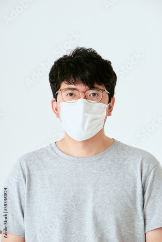 白背景でカメラ目線のマスクと眼鏡を着けた30代の1人の日本人男性の人物の上半身の正面 © chachamal
