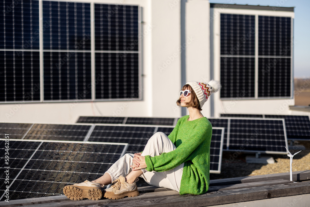 Woman in green sweater and winter hat sitting relaxed on a rooftop of her house with a solar power plant installed on it. Energy independence of household in winter Woman in green sweater and winter hat sitting relaxed on a rooftop of her house with a solar power plant installed on it. Energy independence of household in winter