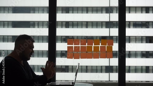 Silhouette of middle aged man with a laptop sitting near a panoramic window on a terrace balcony at home. A man gesticulates with his hands while communicating via video call. Video conference online. photo