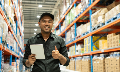 Warehouse worker wearing a hat and black shirt hands holding tablet check stock on tall shelves in warehouse storage. Asian auditor or staff work looking up stocktaking inventory in warehouse store. photo