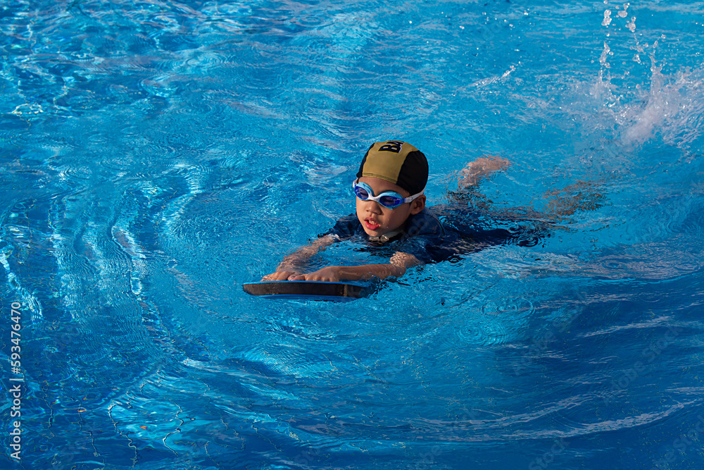 6 years old young boy who a swimmer wearing goggle with dark blue suit and yellow striped black cap. Lonely child warm up with swimming kickboard before practicing to swim in the blue swimming pools.