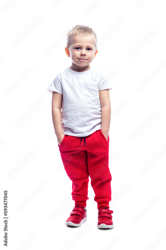 A smiling little blond boy stands with his hands in his pockets. A child in red pants and a white T-shirt. Isolated on white background. Full height. Vertical. A smiling little blond boy stands with his hands in his pockets. A child in red pants and a white T-shirt. Isolated on white background. Full height. Vertical.