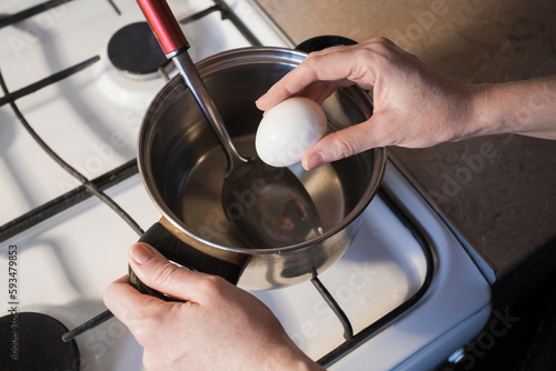 A girl boils egg in a metal pan on a white gas stove in the kitchen. Image for your creative decoration or design.