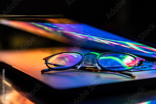 A laptop half closed in the dark with colourful glow and glasses. © puhimec