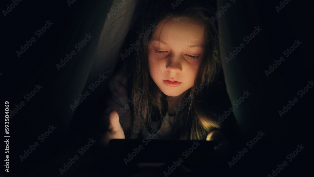 Bedtime phone. Sleepless kid. Internet insomnia. Tired sleepy little girl hiding under blanket using gadget late at night in bed in dark home bedroom. Bedtime phone. Sleepless kid. Internet insomnia. Tired sleepy little girl hiding under blanket using gadget late at night in bed in dark home bedroom.