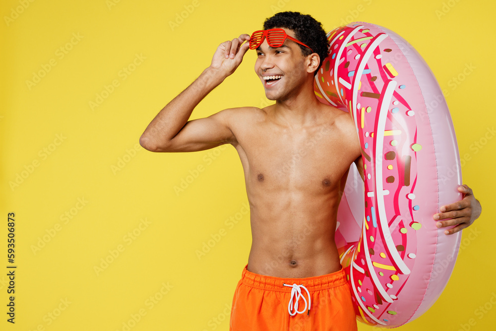 Fun young sexy man wear orange shorts swimsuit take off sunglasses look aside relax near hotel pool hold rubber donut ring isolated on plain yellow background Summer vacation sea rest sun tan concept