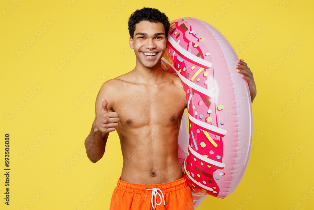 Handsome smiling fun young sexy man wear orange shorts swimsuit relax near hotel pool hold rubber donut ring show thumb up isolated on plain yellow background Summer vacation sea rest sun tan concept