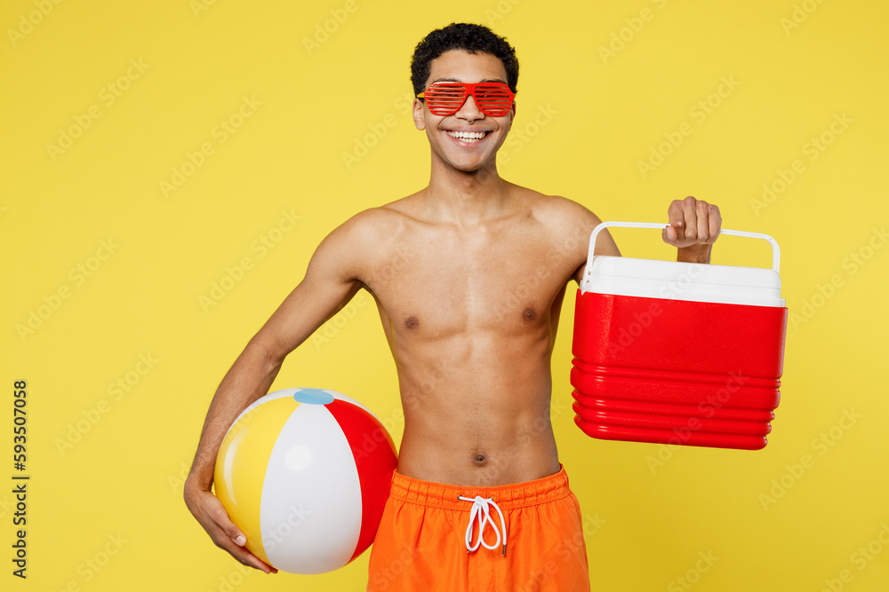 Handsome fun young sexy man wear orange shorts swimsuit glasses hold rubber ball beach refrigerator relax near hotel pool isolated on plain yellow background. Summer vacation sea rest sun tan concept.