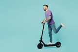 Full body side view caucasian smiling fun happy young man he wear purple t-shirt riding e-scooter raise up leg isolated on plain pastel light blue cyan background studio portrait. Lifestyle concept.