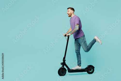 Full body side view caucasian smiling fun happy young man he wear purple t-shirt riding e-scooter raise up leg isolated on plain pastel light blue cyan background studio portrait. Lifestyle concept. photo
