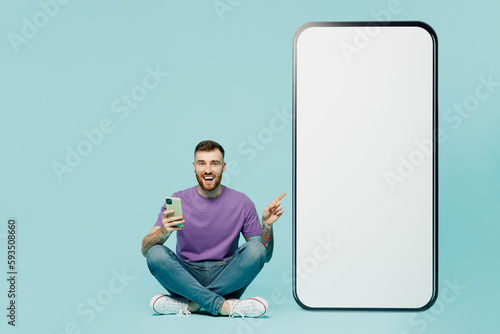 Full body side view fun young man he wears purple t-shirt sit point finger on big huge blank screen mobile cell phone with area use smartphone isolated on plain pastel blue background studio portrait. photo