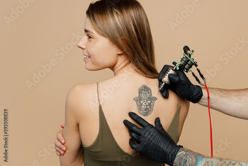 Tattooer master artist hold machine black ink in jar, equipment for making tattoo art on back view smiling happy caucasian woman female body shoulders back isolated on plain beige background studio. photo