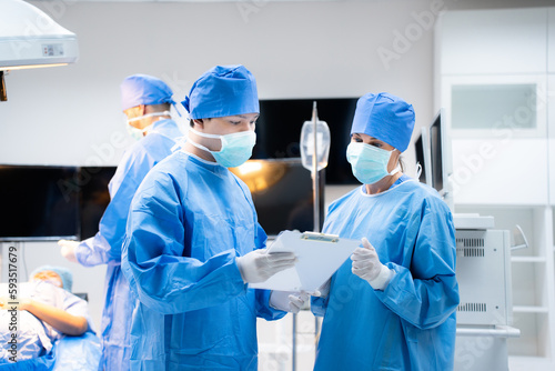 Selective focus front view of Caucasian doctors, man and woman, in surgical uniforms, face masks, and gloves, standing watching the clipboard with medical report in hands in a modern operating room.
