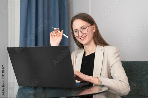 Smart beautiful cute charming attractive young woman with glasses is holding an online meeting via a laptop. Business woman in a light jacket is sitting at a table in a laptop photo