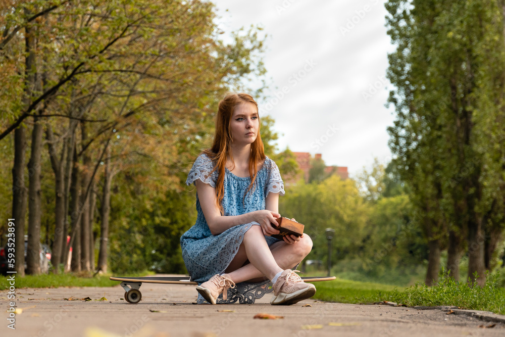 Thoughtful young blond woman sitting on longboard in the middle of park alley, playing music with kalimba and looking away