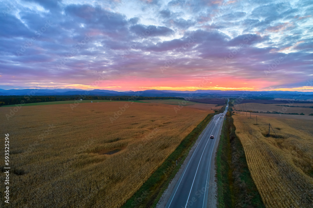 Aerial view of mountain road at sunset in autumn. Top view from drone of road in hills. Beautiful landscape with roadway, trees, green meadows, sky with golden sunlight in fall.