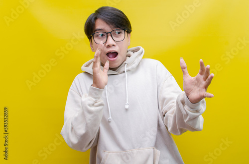 ecstatic, attractive young Asian man wearing beige hoodie shocked, surprised happily opening mouth 