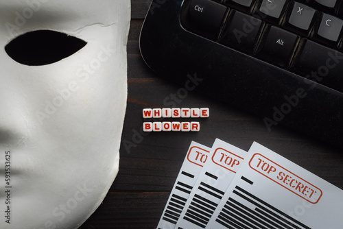 Whistleblowing concept: white mask on a wooden table along with a computer keyboard and classified documents