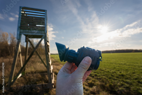 Thermal night vision device in hand against the background of a hunting tower on a field on a sunny day. photo