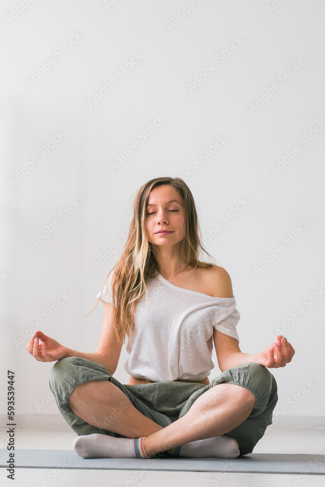 Young healthy beautiful woman practicing yoga at home sitting in lotus pose on yoga mat meditating smiling relaxed with closed eyes - Mindfulness meditation concept