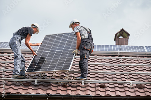 Men technicians lifting photovoltaic solar modul on roof of house. Electricians in helmets mounting solar panel system outdoors. Concept of alternative and renewable energy.