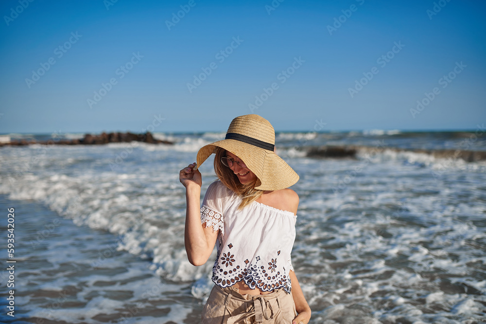 Young pretty girl standing in the waves of the sea and smiling at the camera. Lifestyle girl with glasses and a hat at sea. Pastel colors of clothing.