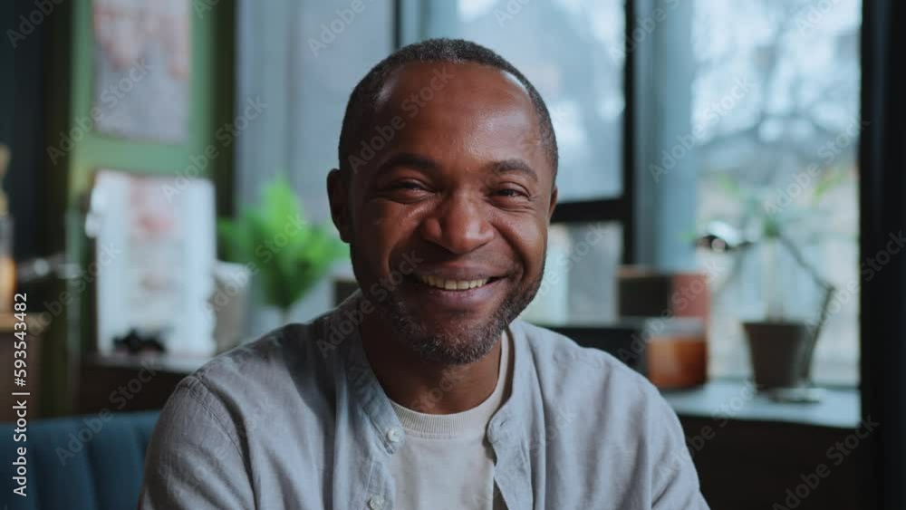 Horizontal shot of smiling handsome African American man in casual outfit looking directly at camera while sitting in front of laptop. Cozy modern interior on blurred background Horizontal shot of smiling handsome African American man in casual outfit looking directly at camera while sitting in front of laptop. Cozy modern interior on blurred background