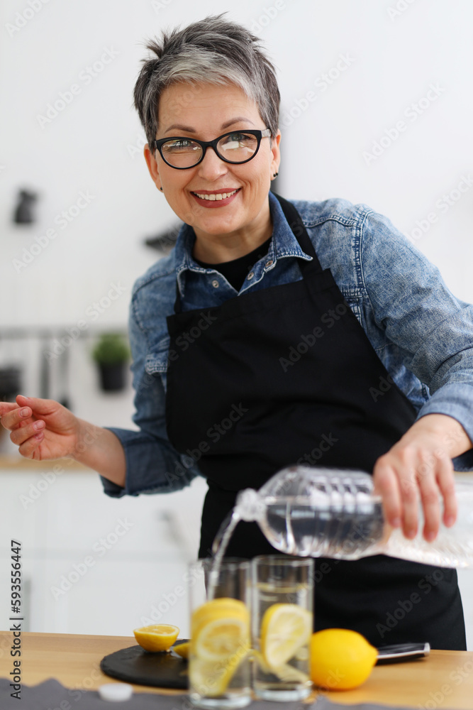Portrait of a positive woman in the kitchen pours water into glasses with lemon. Portrait of a positive woman in the kitchen pours water into glasses with lemon.