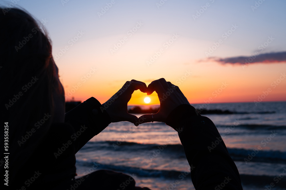 a woman arranges her hands in the shape of a heart against the background of the setting sun over the sea at sunset - obrazy, fototapety, plakaty 