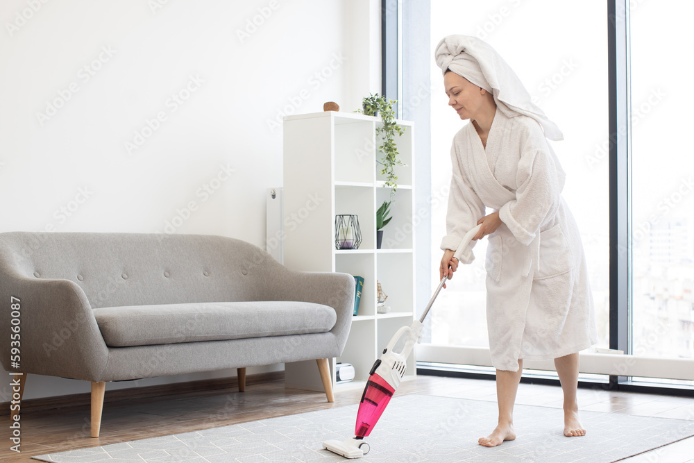 Young woman in bathrobe and towel on head removing dust from soft carpet using cordless vacuum cleaner in spacious living room. Cheerful housewife enjoying housekeeping with modern appliances. Young woman in bathrobe and towel on head removing dust from soft carpet using cordless vacuum cleaner in spacious living room. Cheerful housewife enjoying housekeeping with modern appliances.