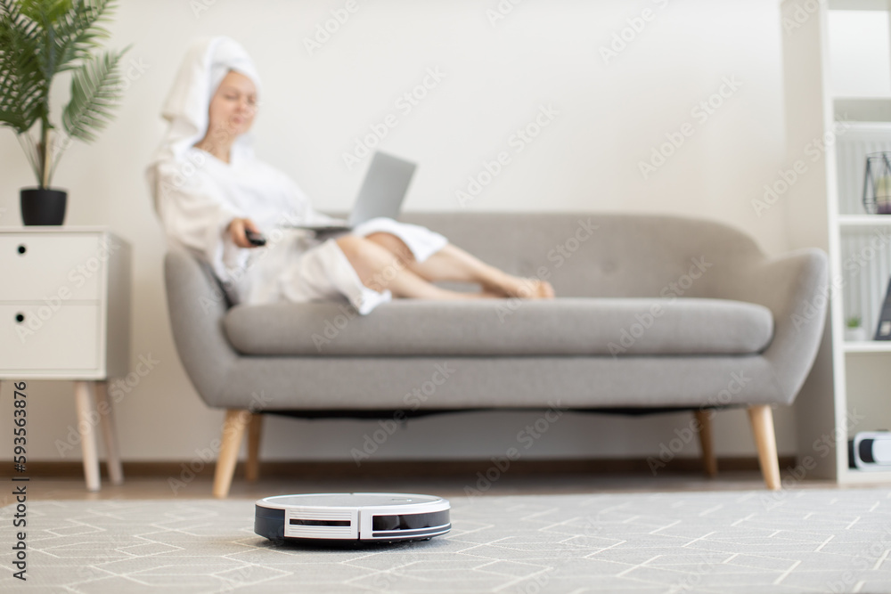 White automatic robot vacuum keeping carpet dust-free while female owner relaxing with portable computer on cozy couch indoors. Wireless tech gadget providing healthy environment in airy apartment. White automatic robot vacuum keeping carpet dust-free while female owner relaxing with portable computer on cozy couch indoors. Wireless tech gadget providing healthy environment in airy apartment.