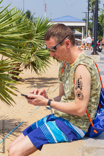 Man sitting on beach hands holding smartphone with freshly applied henna tattoo on forearm