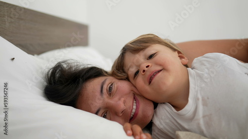 Mother and son laid in bed in embrace looking at camera smiling. Motherhood lifestyle child and mom together