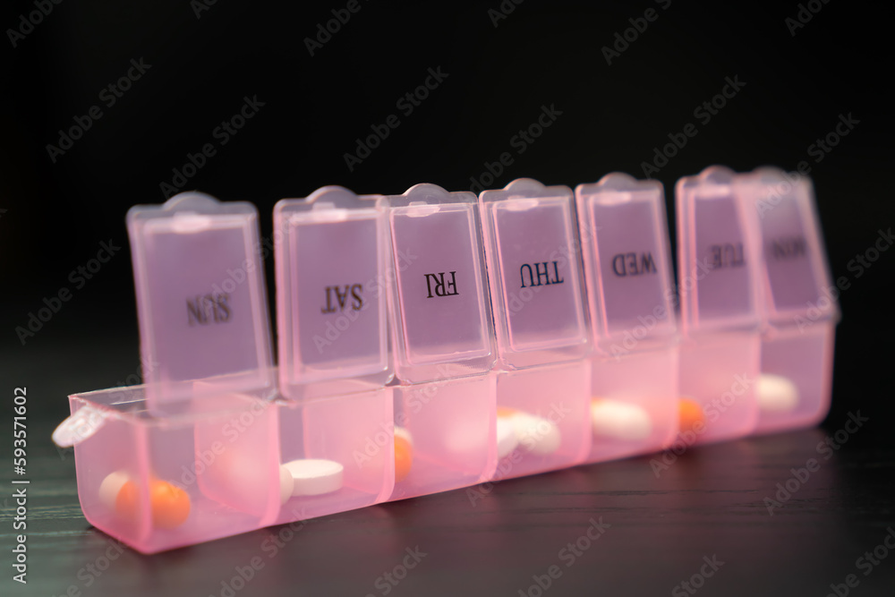 Close-up of a pink pillbox with capsules and tablets on a black table, macro photography. Concept of a healthy lifestyle and medicine Close-up of a pink pillbox with capsules and tablets on a black table, macro photography. Concept of a healthy lifestyle and medicine