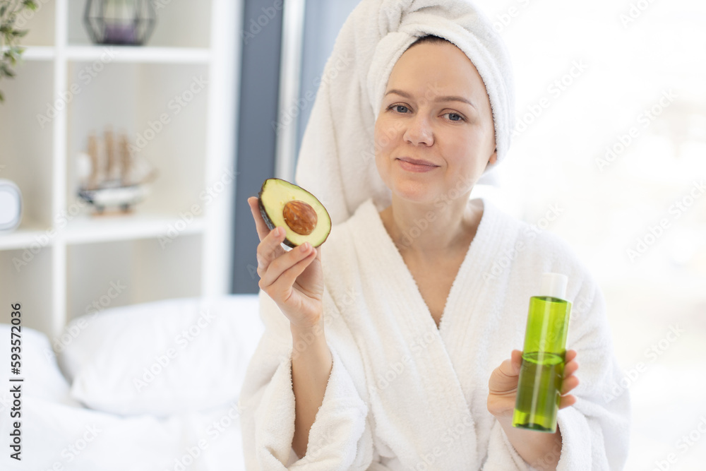 Charming young woman in after-shower outfit enjoying exotic fruit while holding glass vial in bedroom interior. Beautiful female enhancing skin health with avocado facial oil during at-home spa day. Charming young woman in after-shower outfit enjoying exotic fruit while holding glass vial in bedroom interior. Beautiful female enhancing skin health with avocado facial oil during at-home spa day.