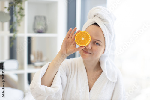 Attractive calm female in white bathrobe and towel on head covering one eye with orange half on blurred background of room interior. Relaxed woman applying citrus rich in vitamin C for beauty care.