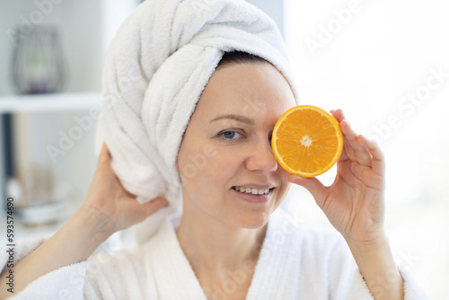 Close-up view of young healthy female wearing soft towel and holding orange fruit piece over her eye on blurred background of bright room. Happy lady achieving natural beauty effect of skin glow.