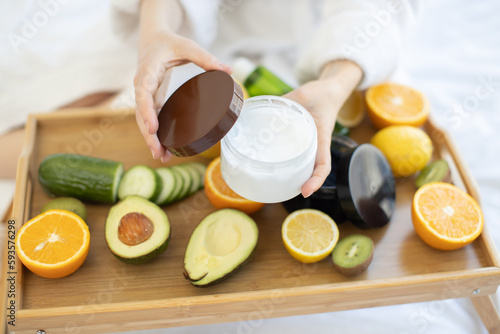 Bild auf Leinwand Young female opening cosmetic product with white creamy texture while sitting on soft blanket near organic beauty remedies on bed tray