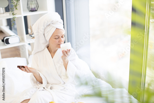 Charming caucasian woman in dressing gown relishing unique scent of body care cream while resting near window in bedroom interior. Good body care routine maintaining natural skin glow effect.