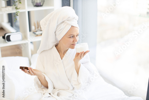 Close-up of lovely carefree lady in towel savouring pleasant fragrance of cosmetic product in hand while pampering herself with skincare procedures. Facial cream moisturizing skin after showering.