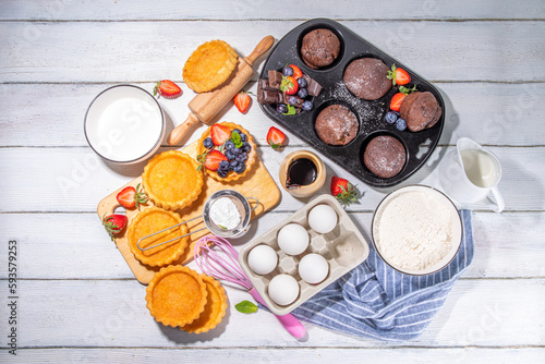 Summer baking background with baked pastry - berry cakes, muffins, mini pie tarts, with cooking baking ingredients, flour, eggs, rolling pin, cream milk, sugar, strawberry, white wooden background