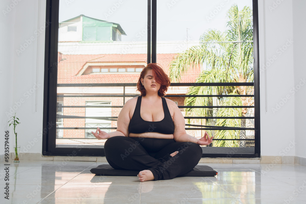 front view of young latin plus size woman sitting doing lotus pose asana indoors. front view of young latin plus size woman sitting doing lotus pose asana indoors.