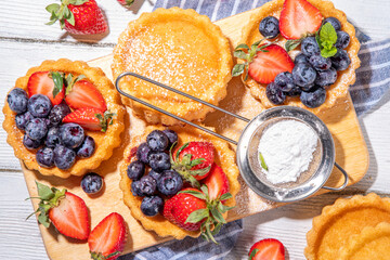 Sponge mini tart cakes with summer berry. Portioned vanilla dessert cups with fresh strawberry, blueberry, raspberry, on white wooden background copy space