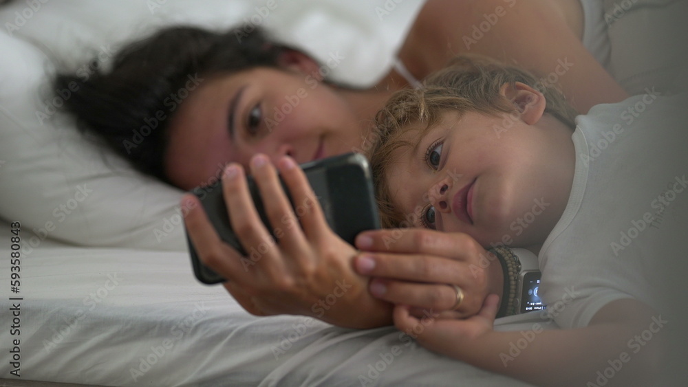 Mom and child looking at phone device together lying in bed. Candid family lifestyle of parent staring at phone screen together - obrazy, fototapety, plakaty 